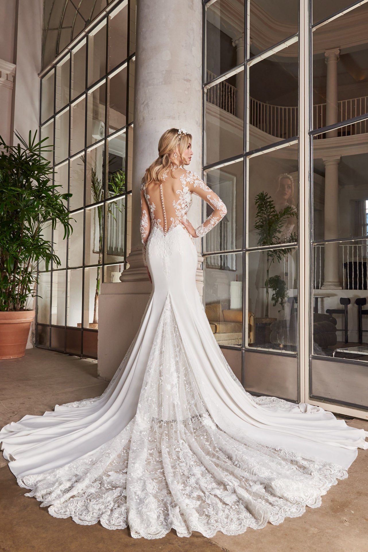 Crepe and lace fishtail gown with high-neck into elegant long lace sleeves. Dramatic illusion back with pearl buttons. Godets of lace embellished with lace appliqué flow into train.