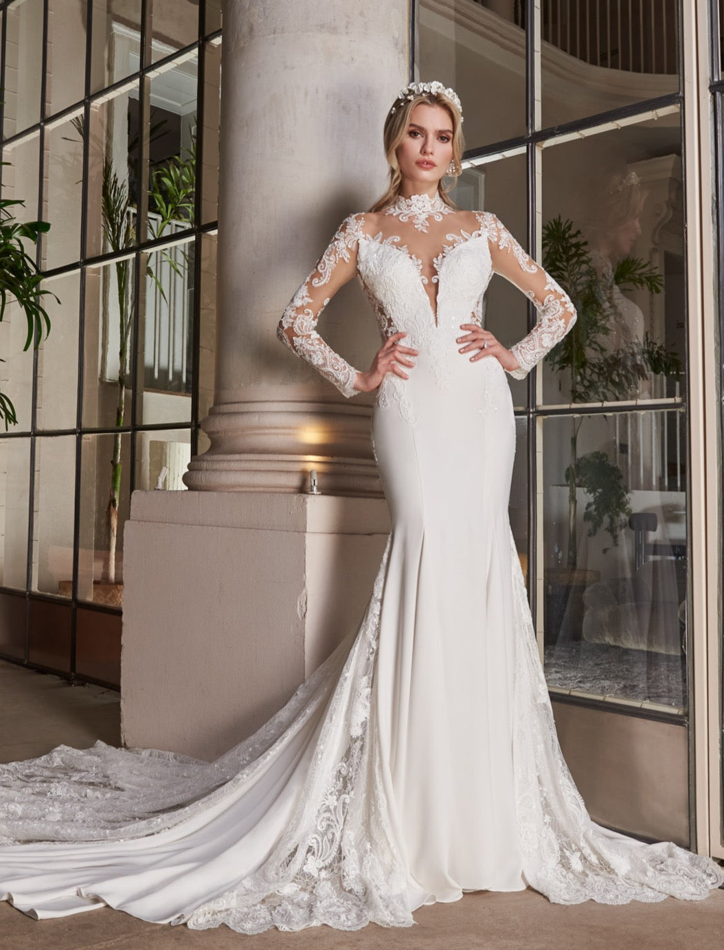 Crepe and lace fishtail gown with high-neck into elegant long lace sleeves. Dramatic illusion back with pearl buttons. Godets of lace embellished with lace appliqué flow into train.