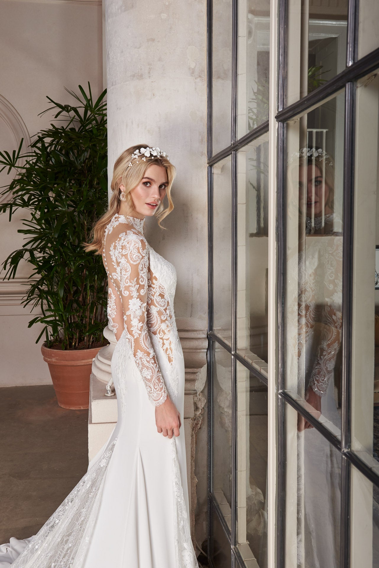 Crepe and lace fishtail gown with high-neck into elegant long lace sleeves. Dramatic illusion back with pearl buttons. Godets of lace embellished with lace appliqué flow into train.