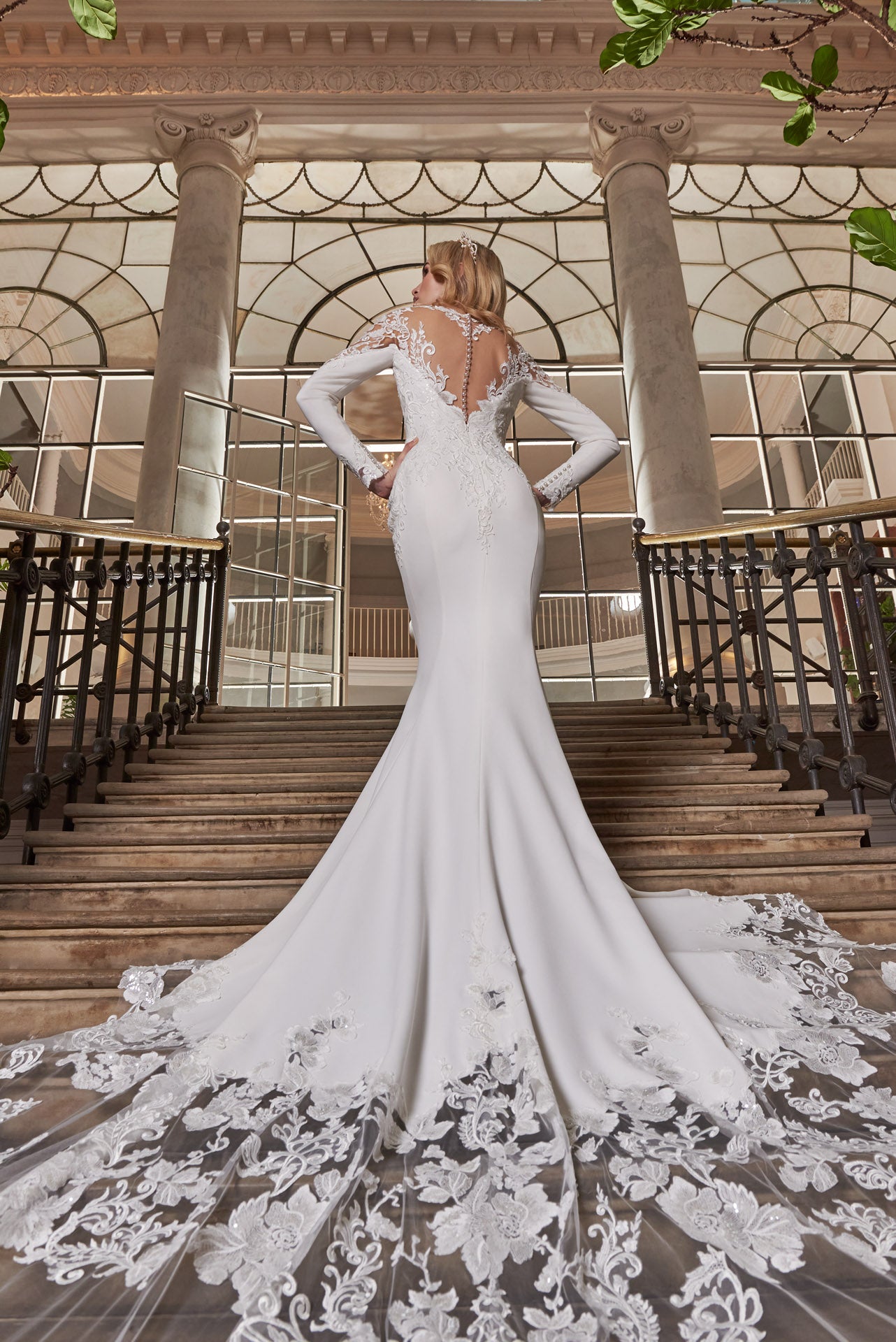 A luxury crepe sculpted dress, lace double tiered train with detailed lace appliqué. A timeless open v-neckline with cut-out sleeves with divine cuff detail. Illusion exposed back